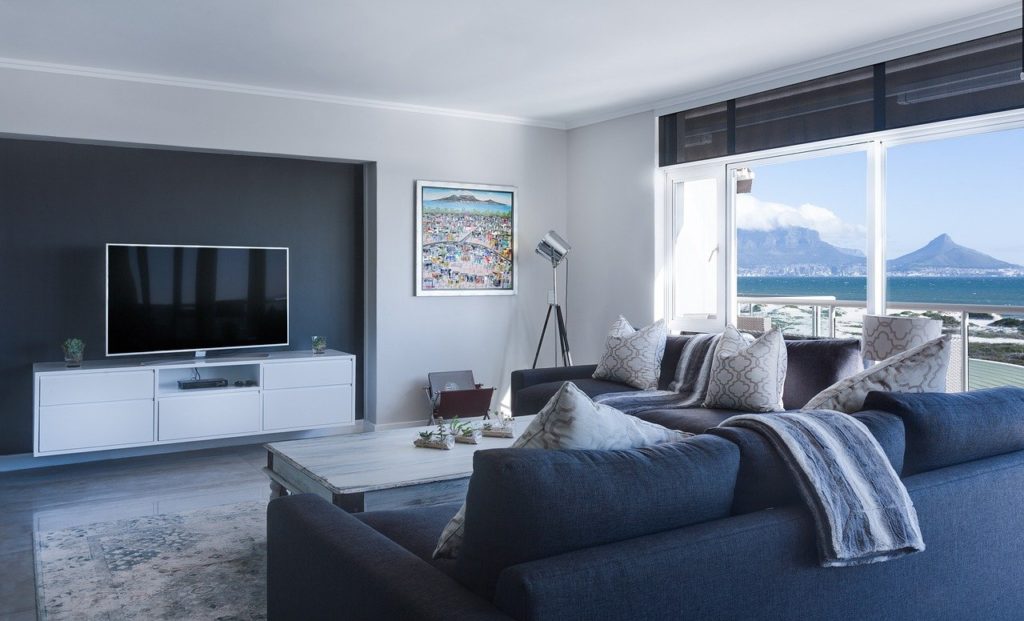 Zona living moderna con divano blu scuro, parete TV bianca e vista panoramica sul mare. Interno luminoso dallo stile elegante e minimalista.