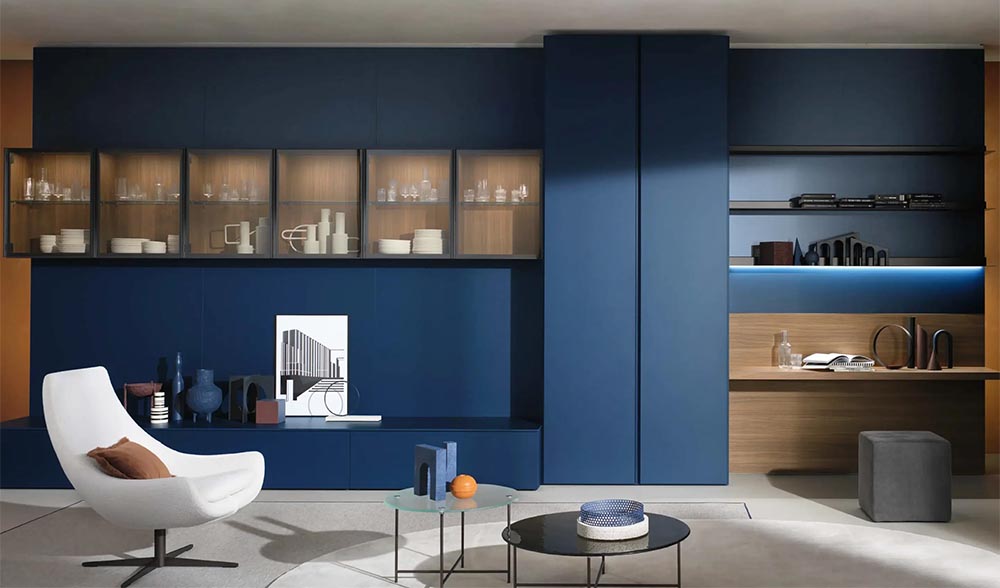 Soggiorno con parete blu opaco, pensili in vetro, postazione studio integrata e arredi design su misura per ambienti raffinati a Milano.