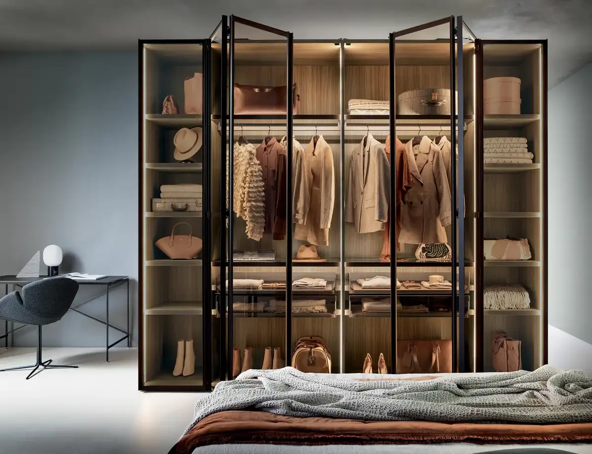 Armadio con ante a libro in vetro trasparente e profili in metallo, illuminazione interna e scaffalature a vista.